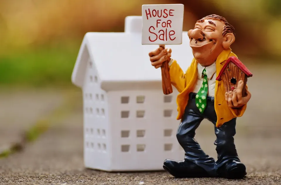Toy figure holding a House for Sale sign beside a small white model house