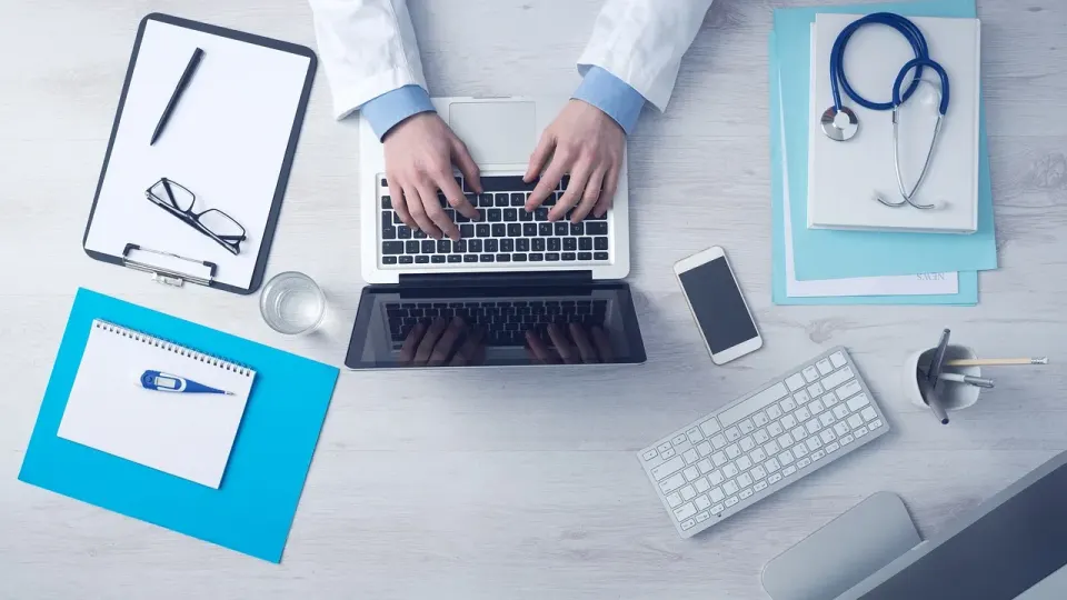 Doctor typing at a laptop on a desk with a stethoscope and clipboard
