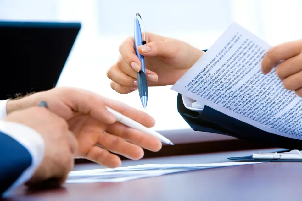 Hand writing notes beside a laptop while reviewing an agreement
