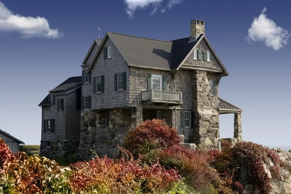Old stone and shingle house with gabled roofs above autumn shrubs